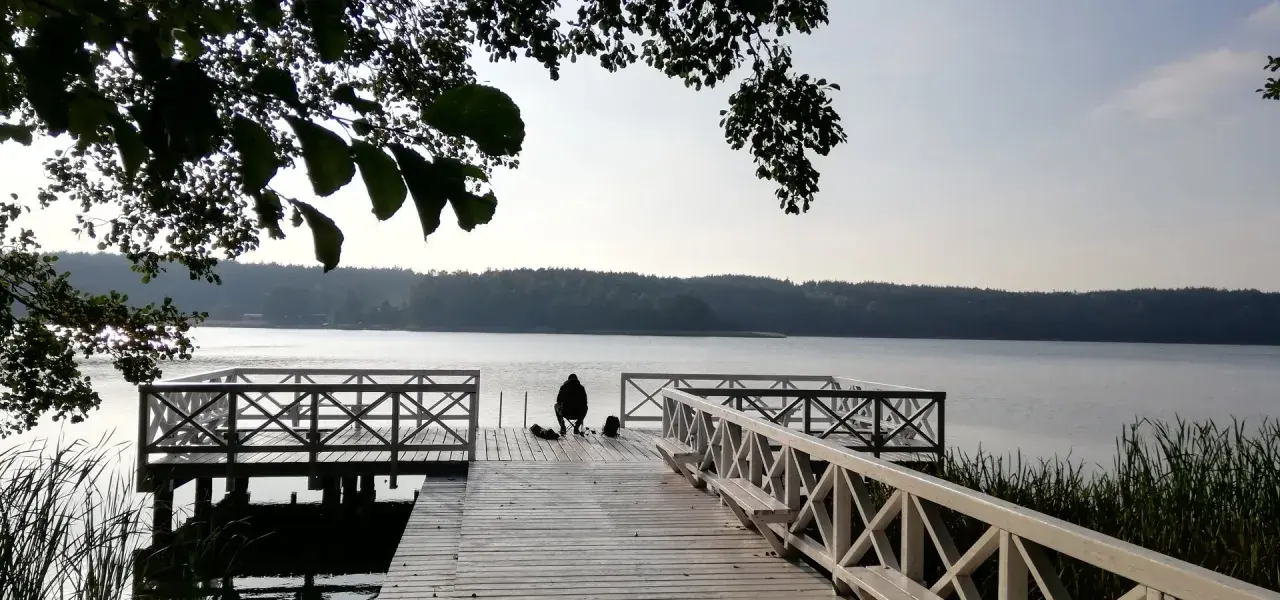 Osoba siedzi na pomoście, przygotowana do wędkowania z noclegiem. Spokojne jezioro i las w tle.