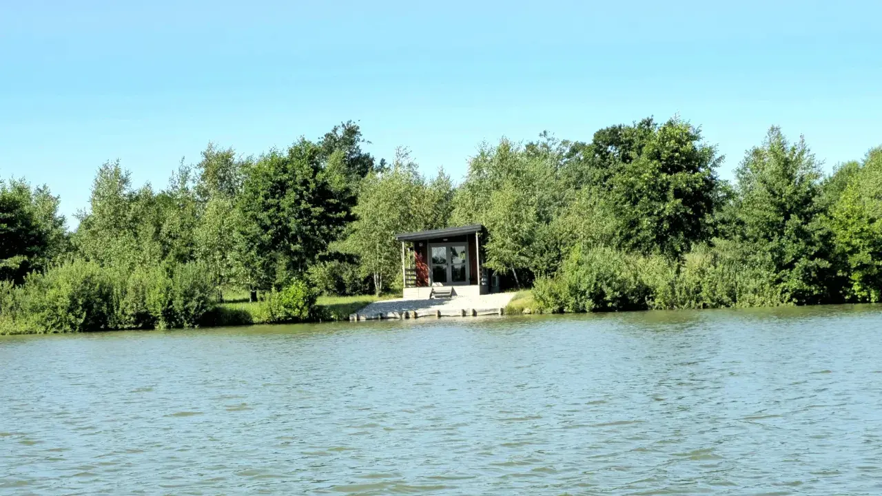 Spokojne wody magnum carp lake otaczają zielone drzewa. Mały domek z tarasem stoi przy brzegu.