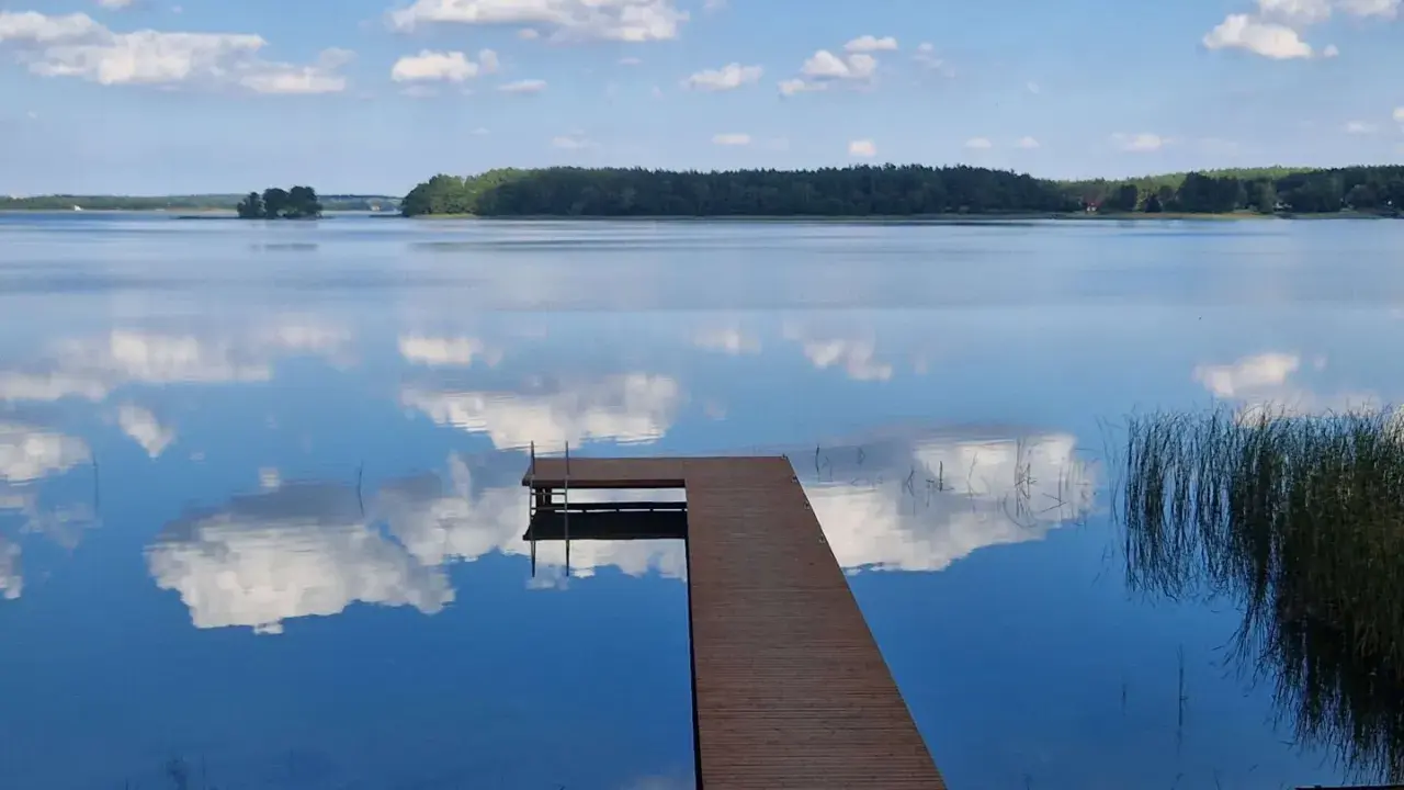 Drewniany pomost na spokojnych jeziorach w mazowieckim, odbijających błękitne niebo z chmurami.