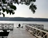 Osoba siedzi na pomoście, przygotowana do wędkowania z noclegiem. Spokojne jezioro i las w tle.