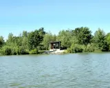Spokojne wody magnum carp lake otaczają zielone drzewa. Mały domek z tarasem stoi przy brzegu.