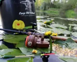 Wędka, kołowrotek i przynęta na liściach lilii wodnych. W tle wiadro z napisem "Carp Old School". Idealne miejsce na łowienie karpi, z widocznymi splawikami karpiowymi.