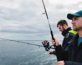 Dwóch wędkarzy na łodzi, uczących się, jak łowić na spinning. W tle morze i pochmurne niebo.