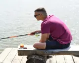 Chłopak z wędką na drewnianym pomoście, zastanawia się, czy wędkarstwo to sport. Słońce odbija się w wodzie.