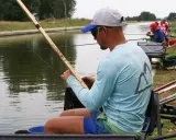 Mężczyzna siedzi nad wodą, trzymając w rękach wędkę bez spławika. W tle inni wędkarze.