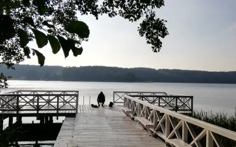 Osoba siedzi na pomoście, przygotowana do wędkowania z noclegiem. Spokojne jezioro i las w tle.