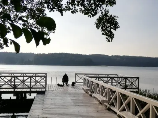 Osoba siedzi na pomoście, przygotowana do wędkowania z noclegiem. Spokojne jezioro i las w tle.