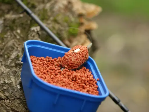 Niebieskie pudełko z zanętą, w której znajduje się pellet ochotka. Na pierwszym planie widać haczyk z przynętą.