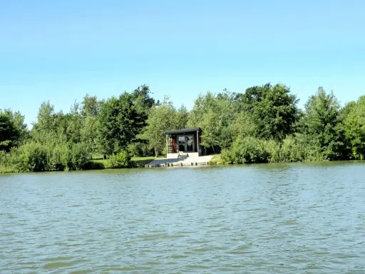 Spokojne wody magnum carp lake otaczają zielone drzewa. Mały domek z tarasem stoi przy brzegu.