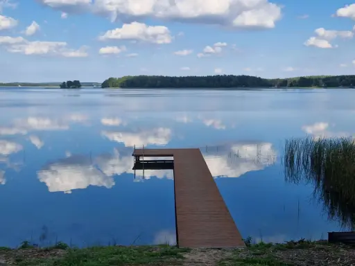 Drewniany pomost na spokojnych jeziorach w mazowieckim, odbijających błękitne niebo z chmurami.