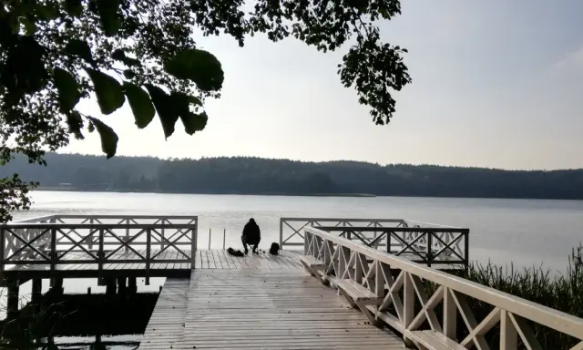 Osoba siedzi na pomoście, przygotowana do wędkowania z noclegiem. Spokojne jezioro i las w tle.