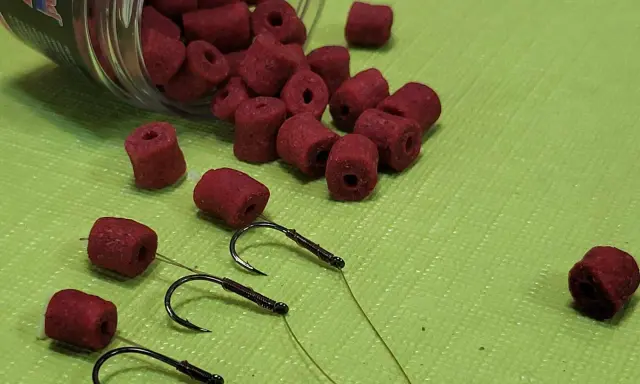 Czerwone przynęty na haczykach, obok rozsypanych z pojemnika. Różne rozmiary haczyków i przynęt.