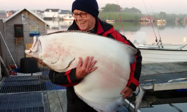 Szczęśliwy wędkarz trzyma ogromnego halibuta. Zapach halibut na jakie ryby - to pytanie zadaje sobie każdy, kto zobaczy ten okaz!