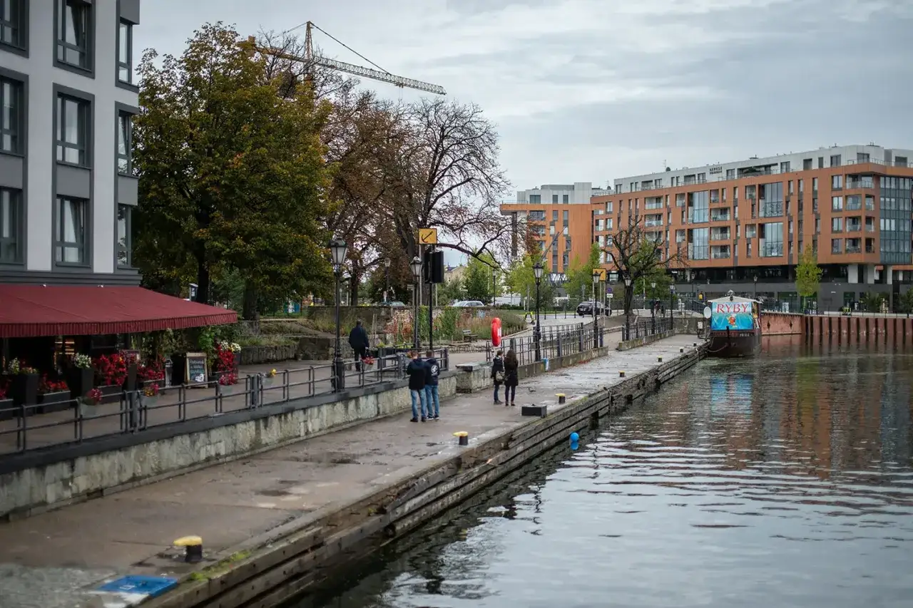 Spacerowicze na nabrzeżu, budynki mieszkalne i łódź z napisem
